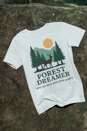 Beige Forest Dreamer T-Shirt with deer and pine trees, laid flat on grass