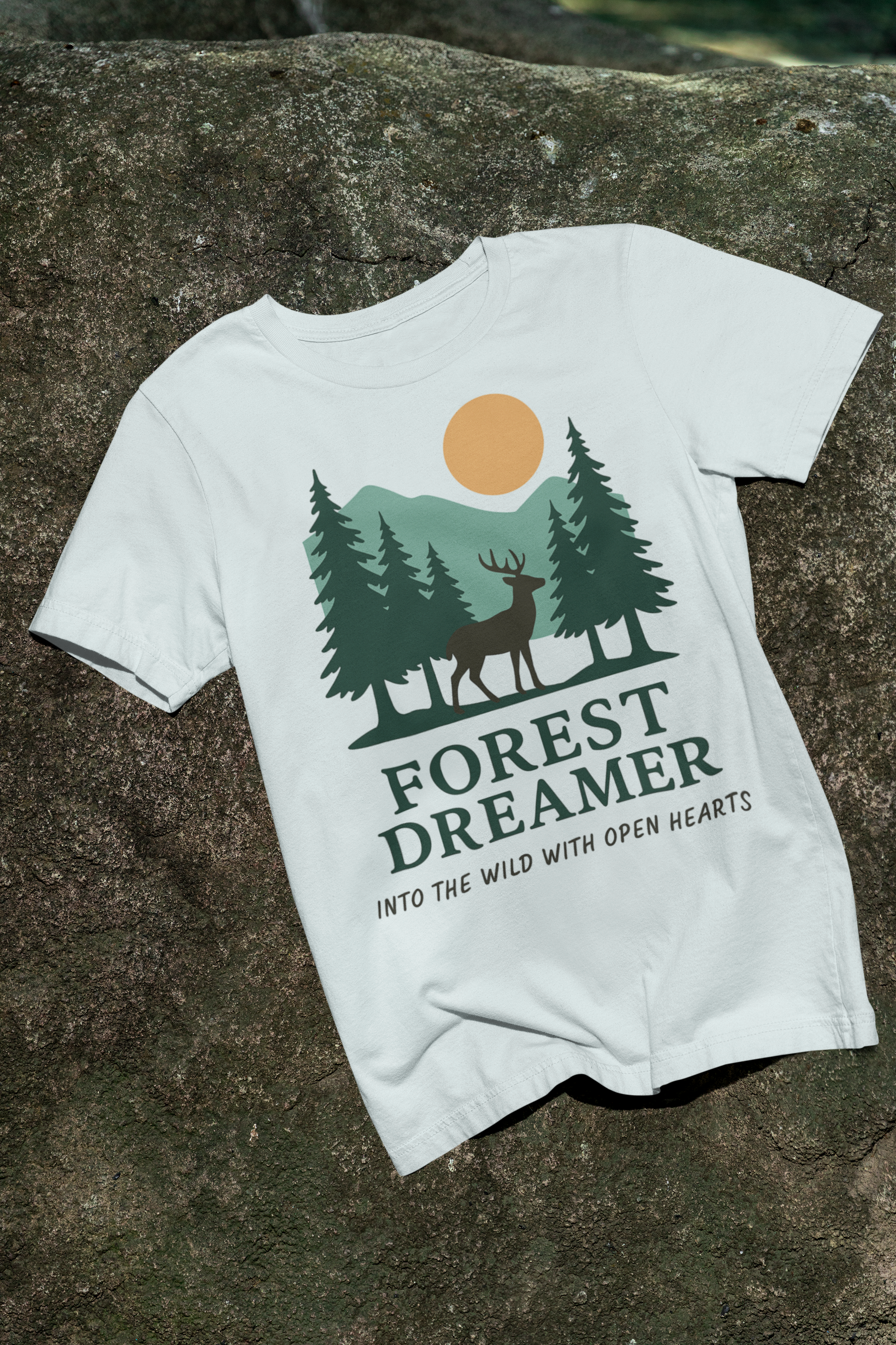Beige Forest Dreamer T-Shirt with deer and pine trees, laid flat on grass