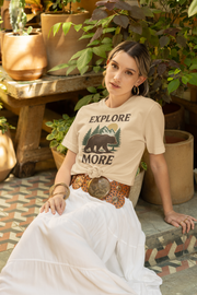 Woman sitting outdoors wearing an Explore More t-shirt, surrounded by potted plants and natural earthy decor.