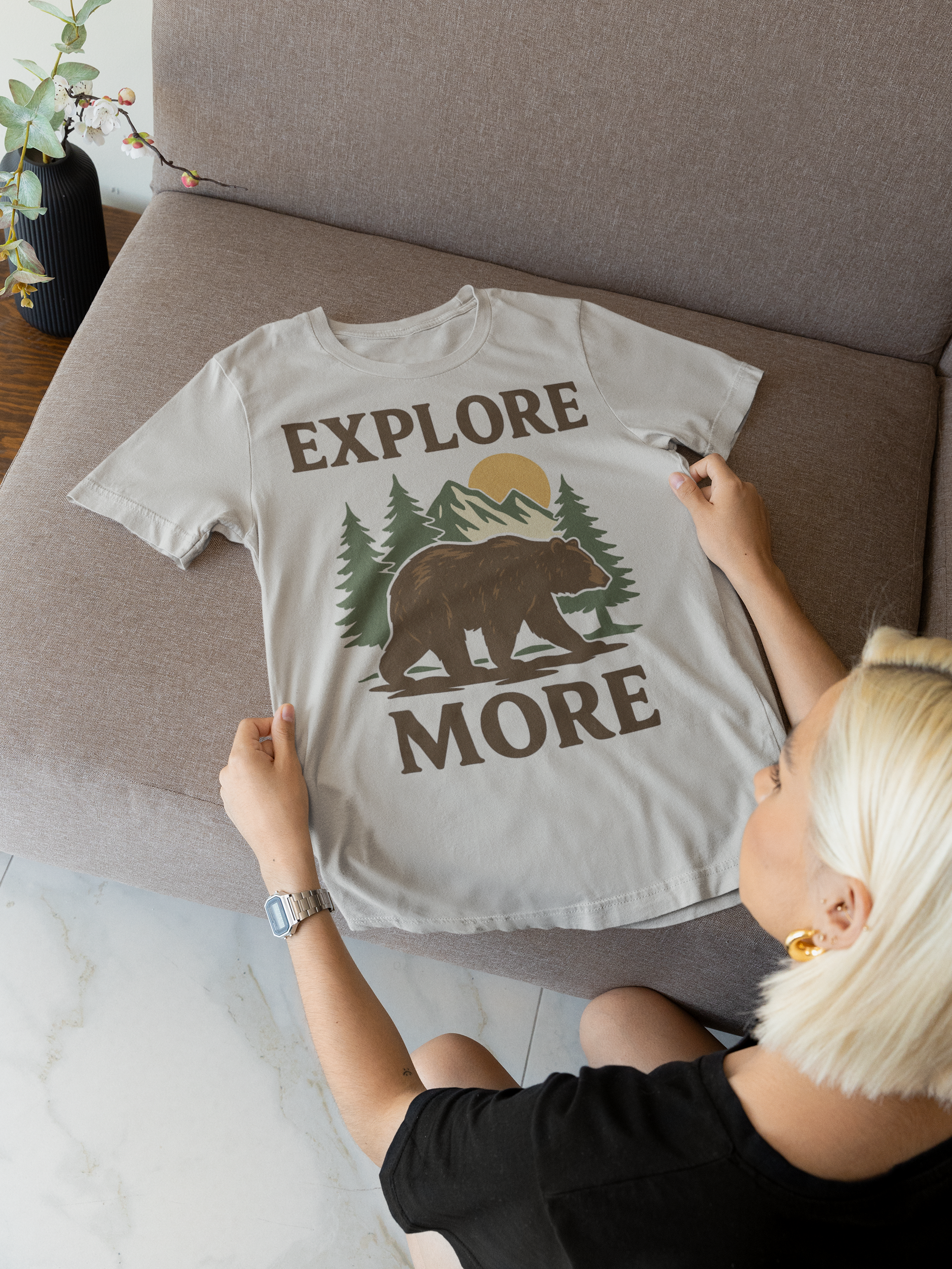 Blonde woman holding a gray Explore More t-shirt with bear and mountain design while sitting on a couch.