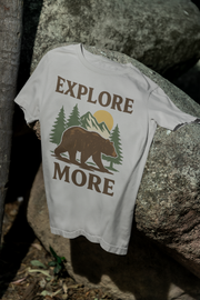 White Explore More t-shirt with bear and forest design hanging on a rock in a natural outdoor setting.