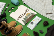 Flat lay of folded Explore More t-shirt with bear and mountain design, surrounded by hiking boots, compass, map, sunglasses, and camping gear on a green background.