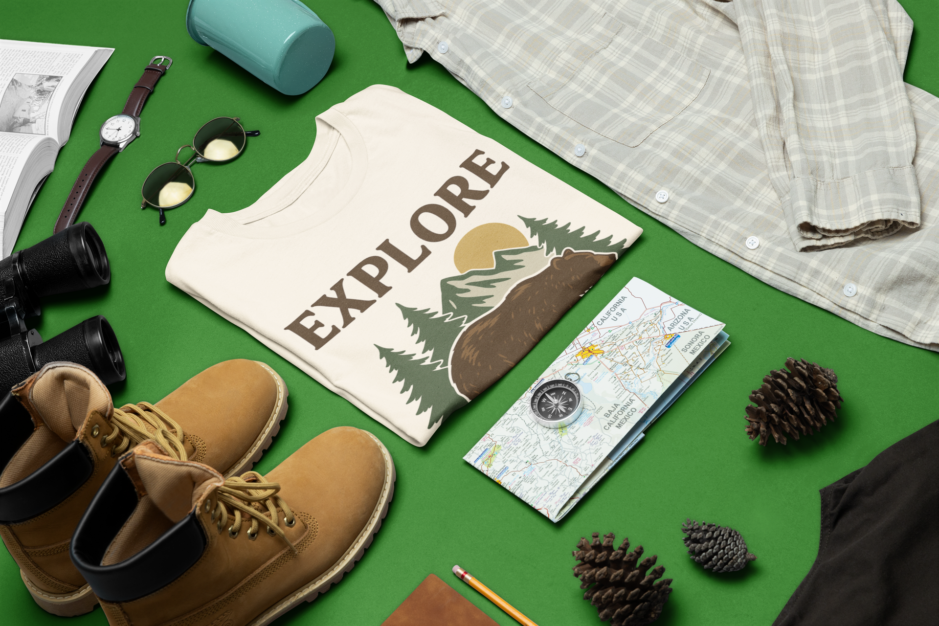 Flat lay of folded Explore More t-shirt with bear and mountain design, surrounded by hiking boots, compass, map, sunglasses, and camping gear on a green background.