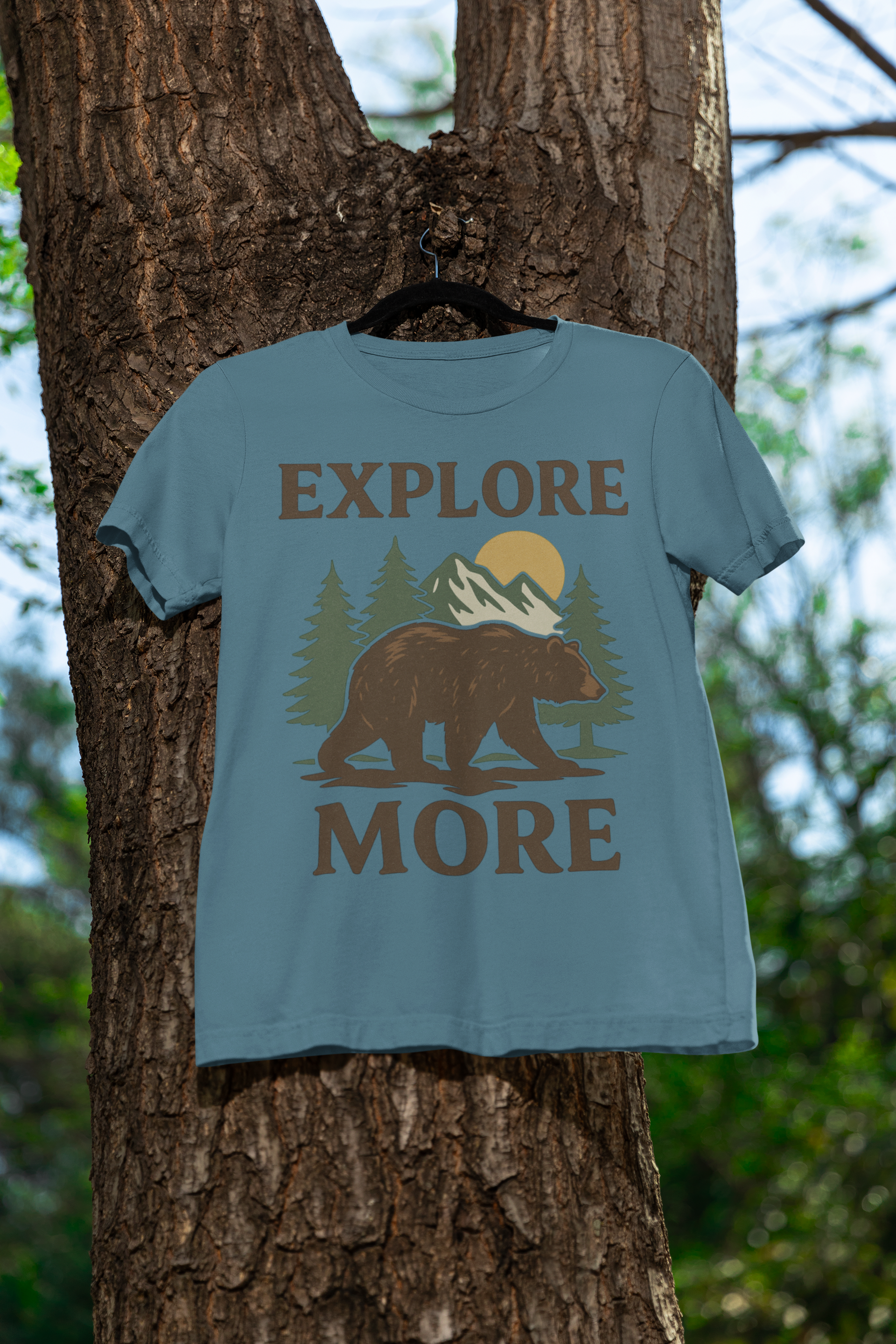 Light blue explore more t-shirt bear and mountain design, hanging on a tree in the forest. Perfect outdoor scene to showcase adventure-inspired apparel.